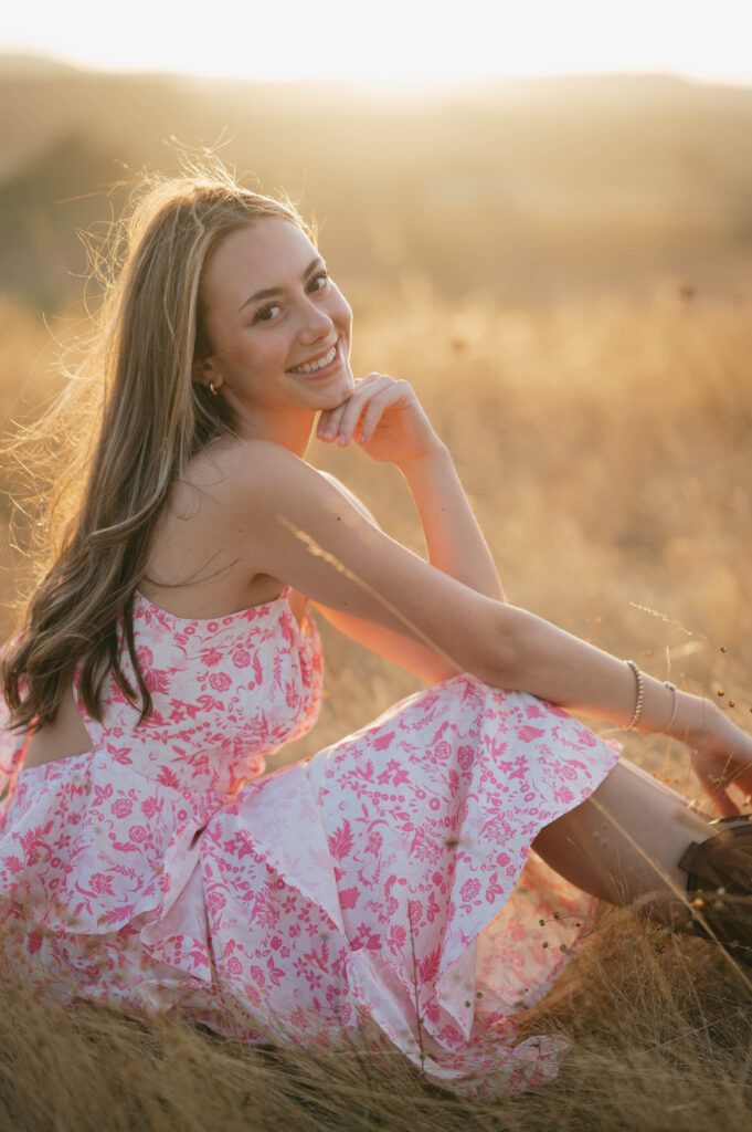 high school senior in pink dress sitting in a grassy field.